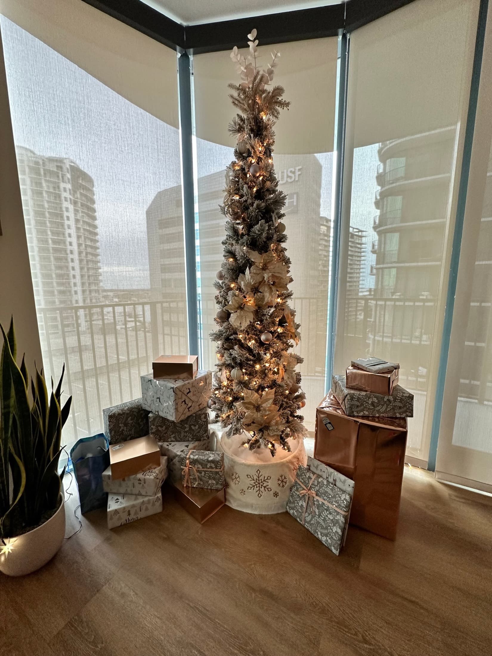 Slim champagne Christmas tree with wrapped gifts beside tall city windows.